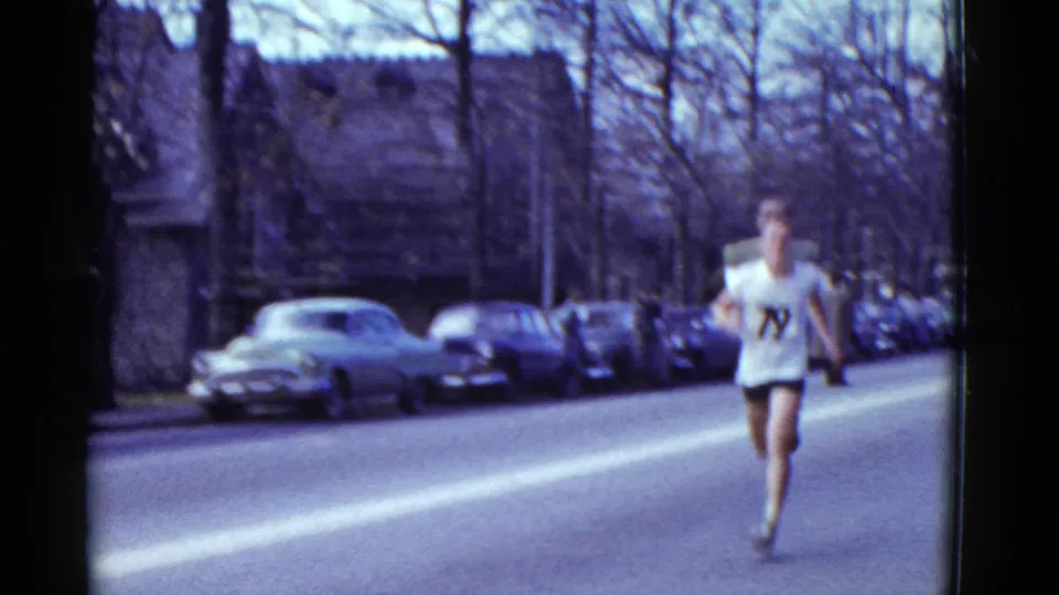Distant 1951 Boston race frame with a male runner approaching along a residential road bordered by parked cars and leafless trees.