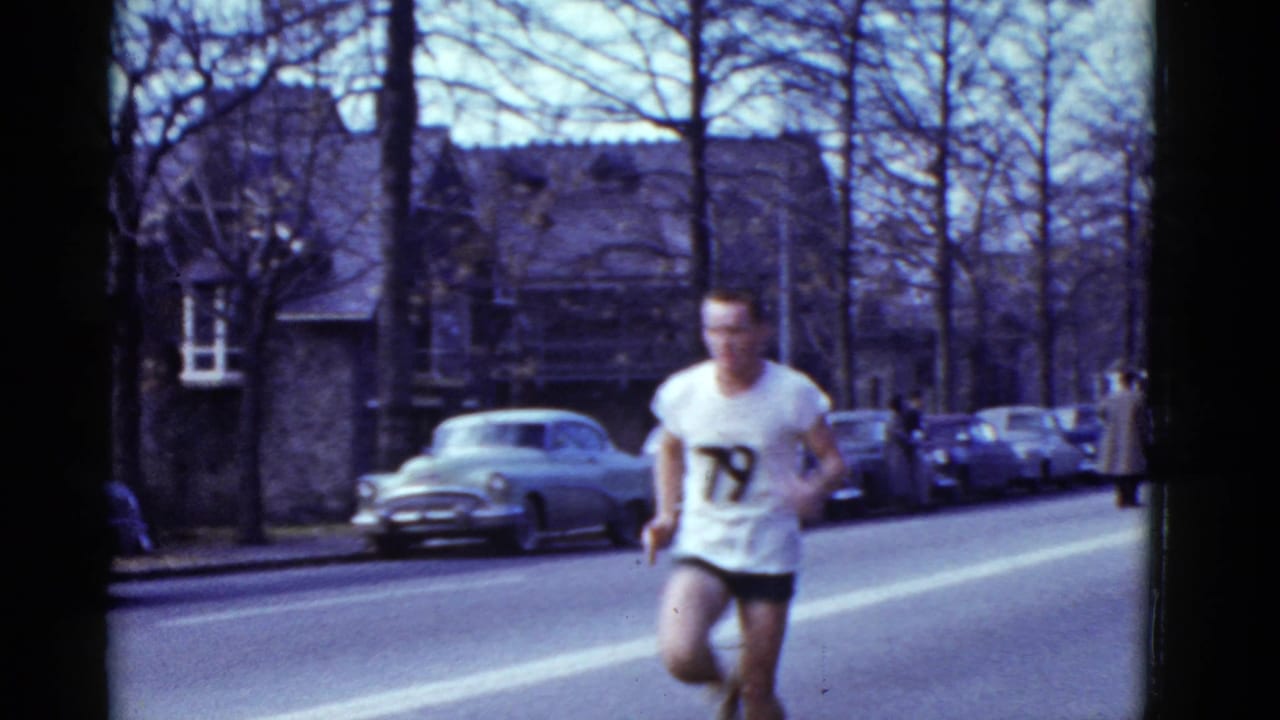 Runner in a white numbered jersey passes parked cars on a tree-lined street.