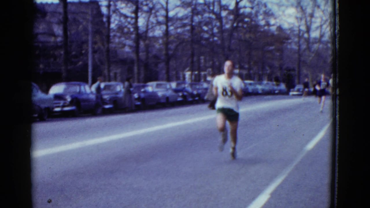 Runner wearing number 83 approaches on a city street lined with parked cars.
