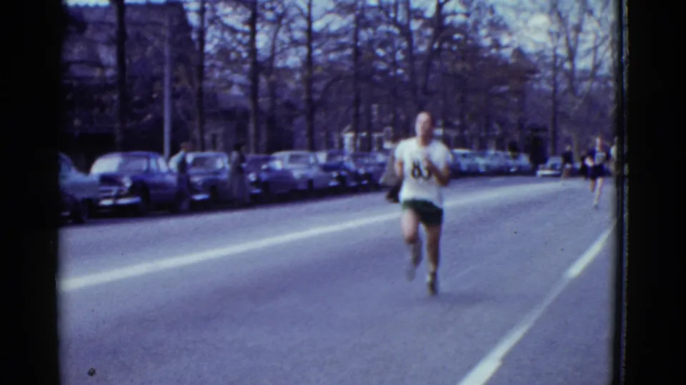 In Boston road-race footage from 1951, a runner in a white number 83 shirt and green shorts moves toward the camera past parked cars.