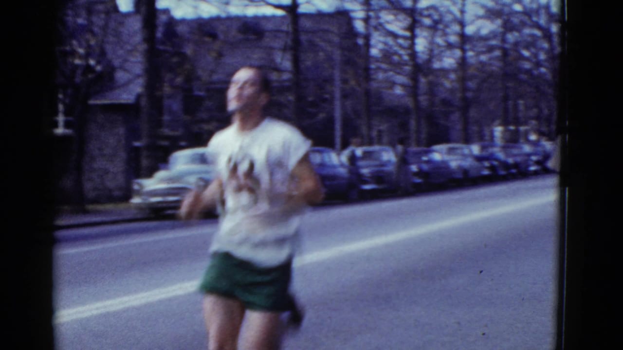 Runner number 83 passes close to the camera with parked cars behind him.