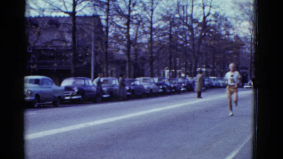 A runner in darker clothing approaches from farther down the Boston street, framed by parked cars, bare trees, and spectators.