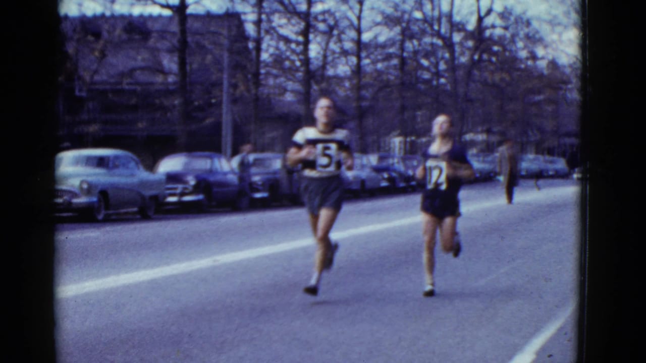 Two numbered runners move down the street with parked cars behind them.