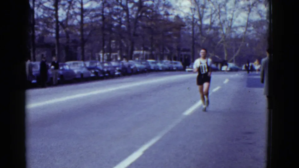 In 1951 Boston, a male runner wearing a numbered bib approaches the camera on a broad street with parked cars and onlookers along the curb.