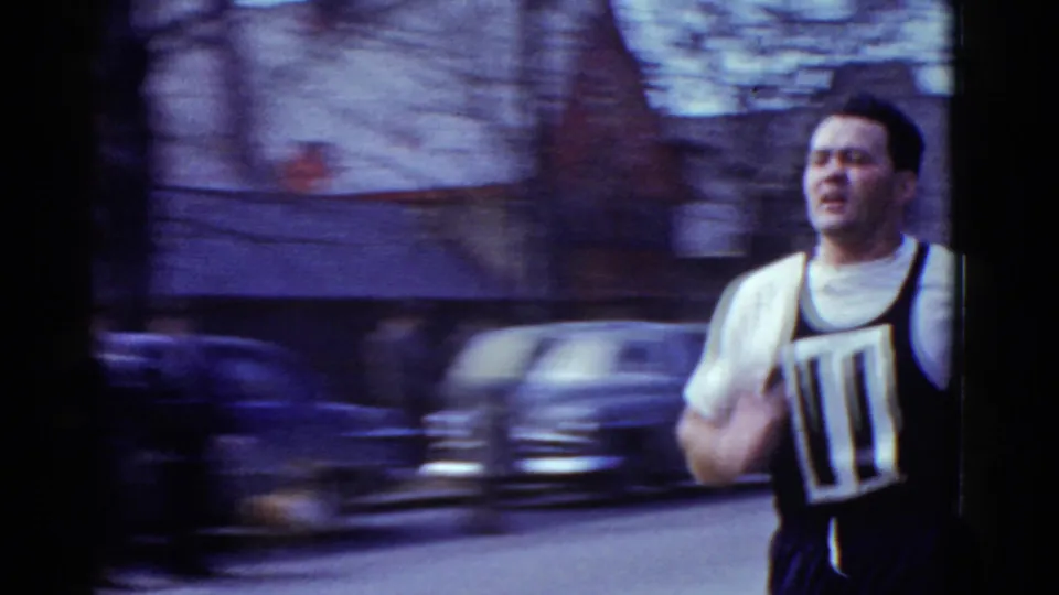 A motion-blurred close view shows the bibbed runner passing the home-movie camera on the Boston street, with cars and buildings behind him.