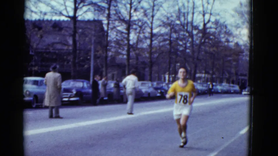 A runner in a yellow top with bib number 78 approaches on the Boston street, while spectators and parked period cars line the roadside.