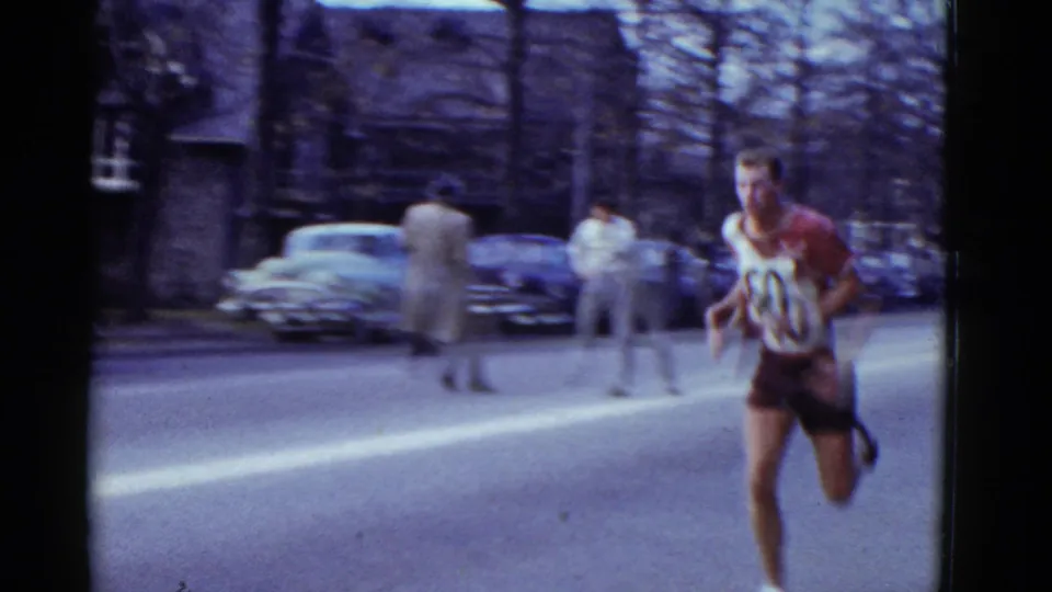 A male runner wearing a red and white uniform with bib 60 runs past parked cars and sidewalk spectators in Boston.