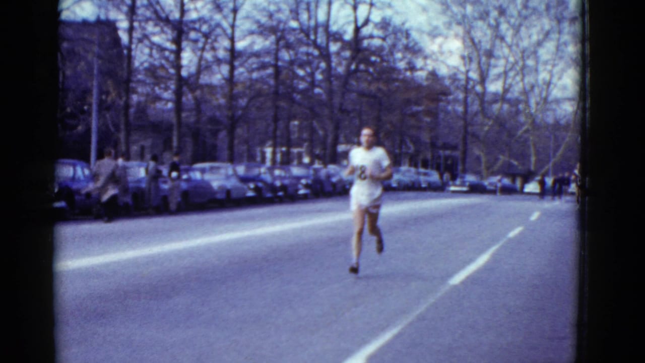 In supported 1951 Boston footage, a man in a white athletic shirt runs toward the camera on a street lined with parked cars.