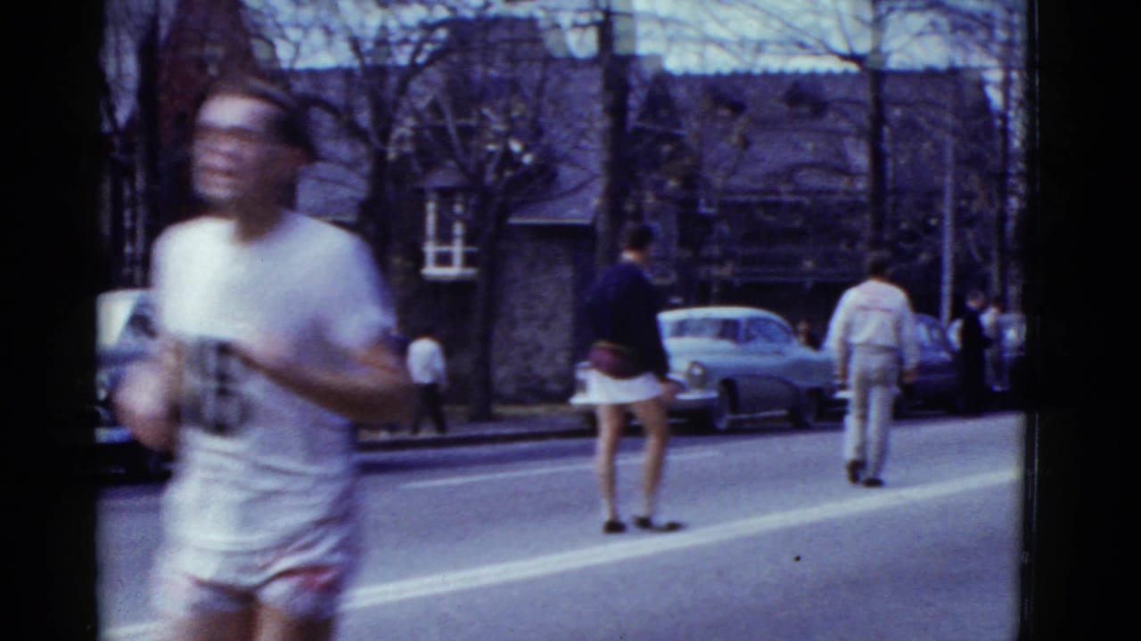 A runner in white blurs past the foreground while another athlete and sidewalk viewers remain visible on the Boston street.