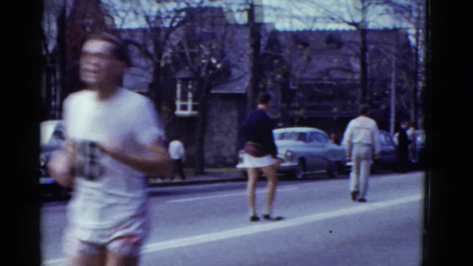 A runner in white blurs past the foreground while another athlete and sidewalk viewers remain visible on the Boston street.