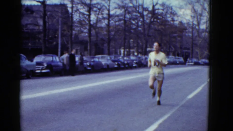A different numbered runner appears to approach the camera on the same Boston street, with spectators and parked cars along the curb.