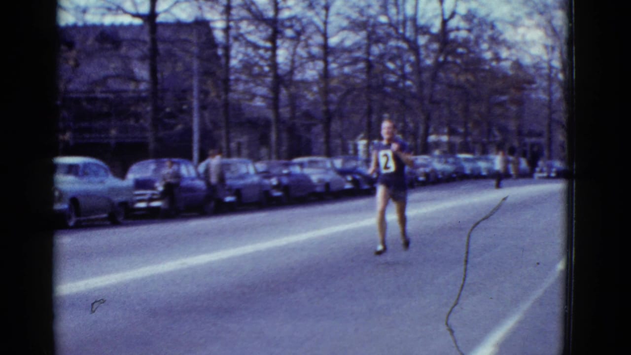 A numbered runner moves toward the camera on the Boston street race route, with parked cars and winter-bare trees behind him.