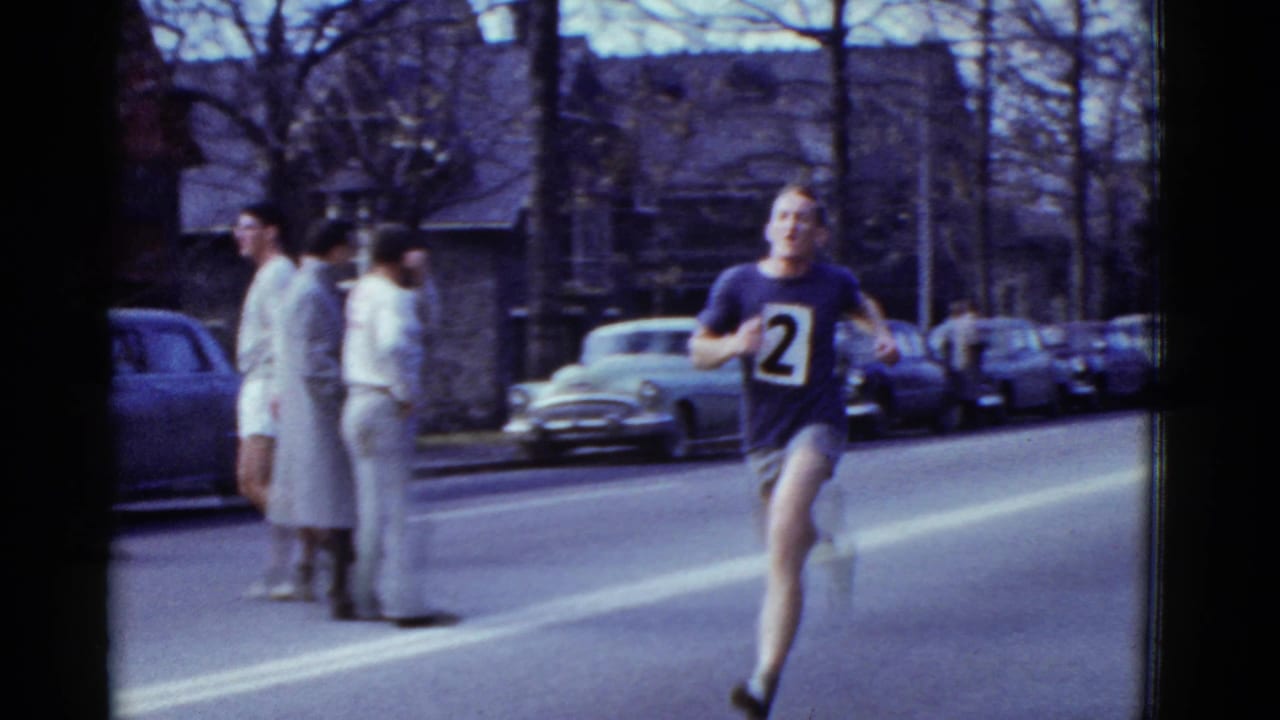 Runner number 2 passes close to the camera in the 1951 Boston street race, with two spectators and parked cars at the curb.