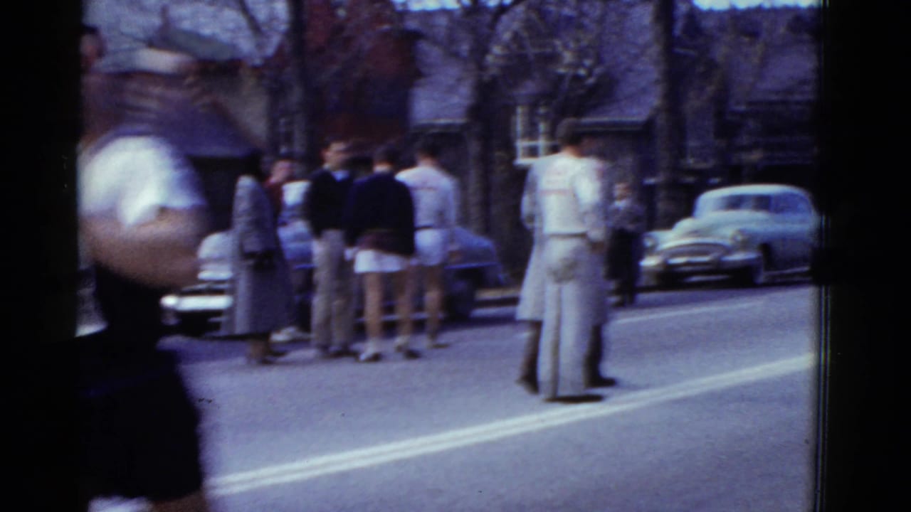 A close passing runner creates motion blur as spectators in period clothing stand near parked cars on the Boston race route.