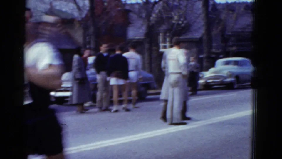 A close passing runner creates motion blur as spectators in period clothing stand near parked cars on the Boston race route.