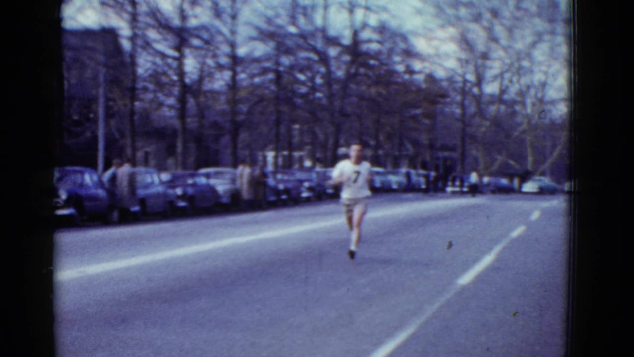A runner in light clothing moves along a broad Boston street in 1951, framed by parked cars, bare trees, and a long road perspective.