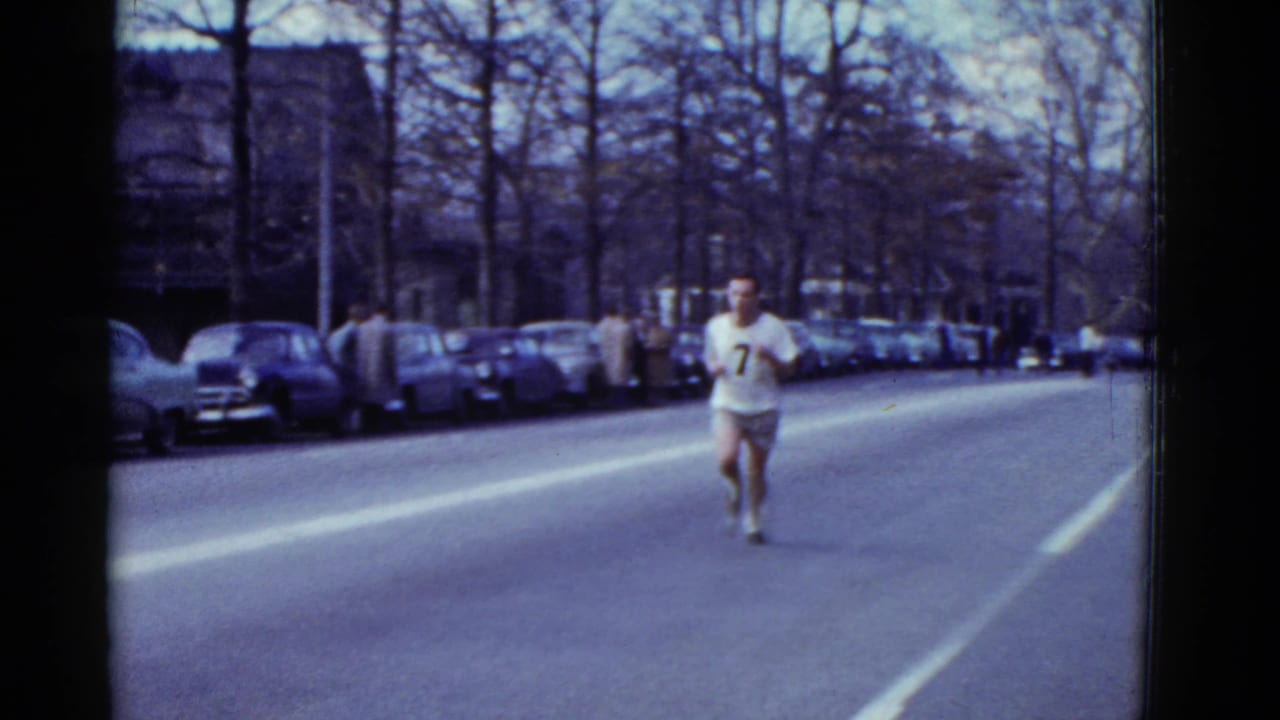 Male runner wearing number 7 approaches on a wide street lined with parked cars and bare trees.