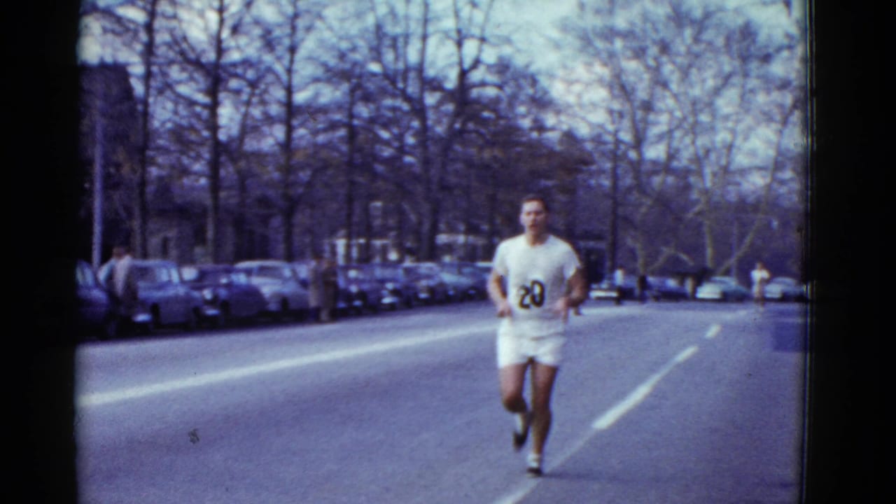 A male runner with number 20 on a white uniform approaches on the road, with parked cars, bare trees, and a few roadside figures visible.