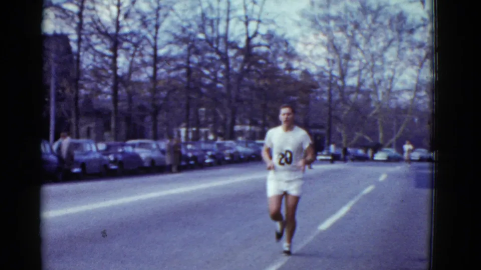 A male runner with number 20 on a white uniform approaches on the road, with parked cars, bare trees, and a few roadside figures visible.
