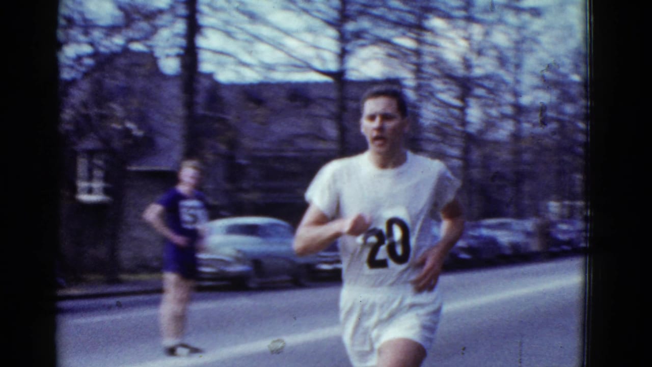 Runner number 20 fills the foreground as a second runner in a dark uniform appears behind him on the street in the supplied Boston clip.