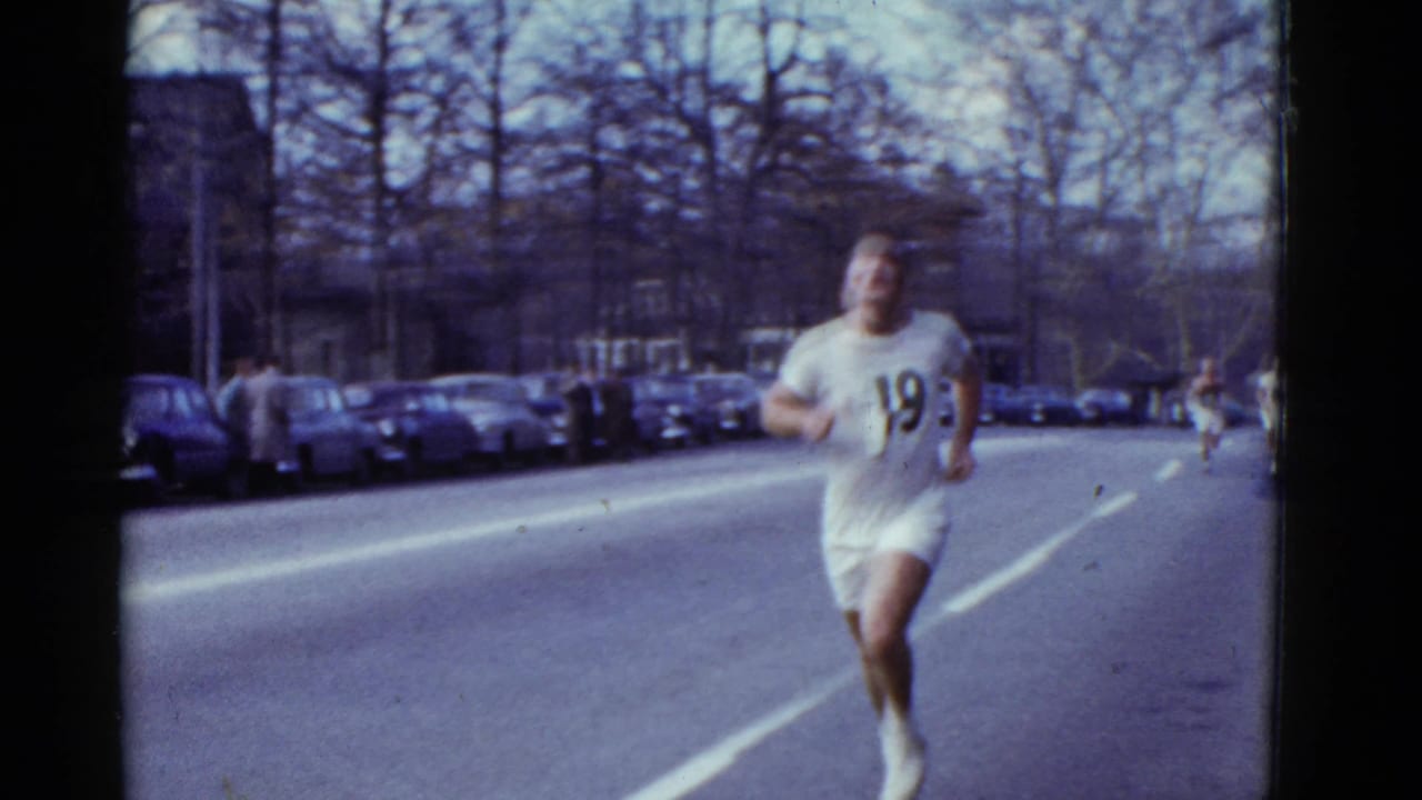Runner number 19 approaches the camera on a Boston street lined with parked mid-century cars.