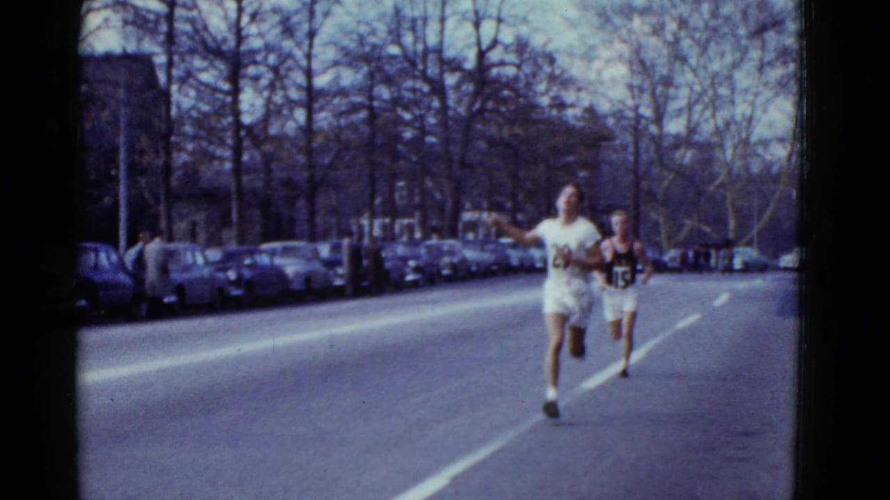 Two runners in numbered uniforms approach on a 1951 Boston street with spectators and parked cars nearby.