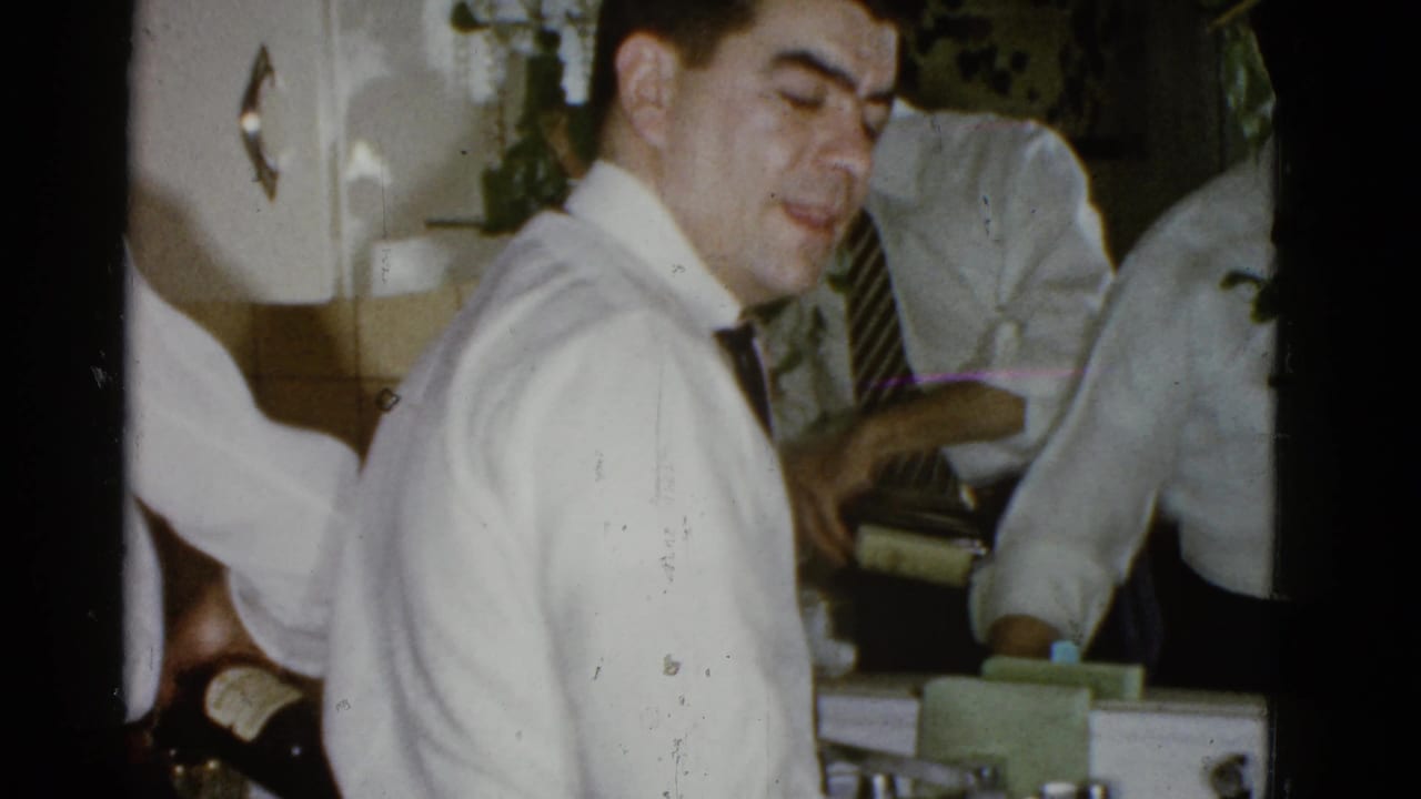 Man in a white shirt and dark tie stands by a kitchen sink with other men reflected nearby.