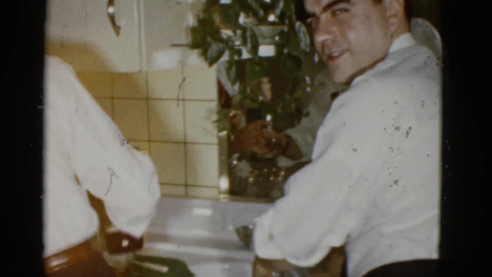 A man in a white shirt turns toward the camera in a 1951 Boston kitchen, with a bottle and sink visible beside him.