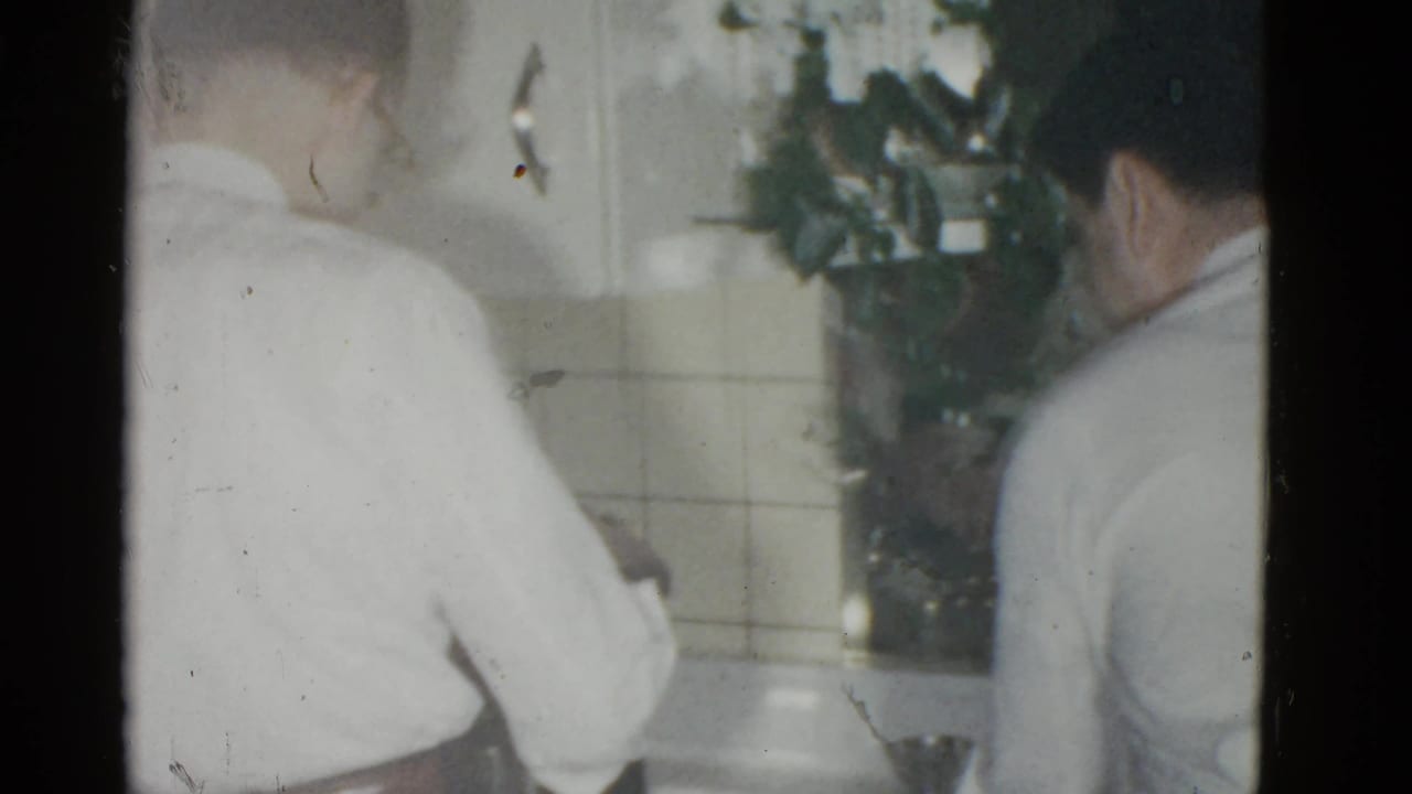 Two men in white shirts stand near a tiled kitchen counter, partly obscured by film exposure and framing.