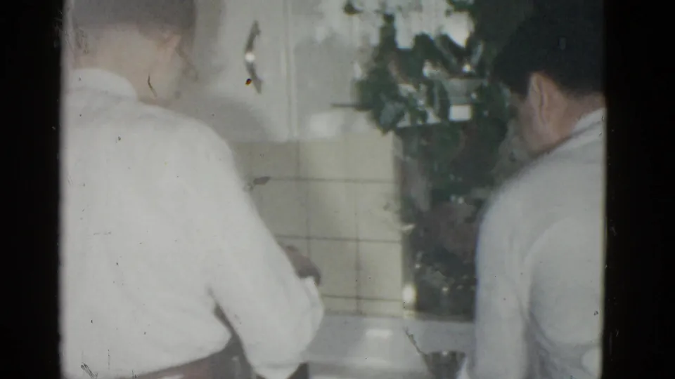 In a 1951 Boston home-movie frame, two adult men stand in a kitchen with beige tile and cabinets, partly softened by 8mm exposure.