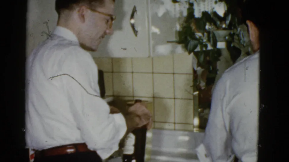 The man in glasses looks down as he handles a bottle or glass in the Boston kitchen, with another adult man partly visible nearby.