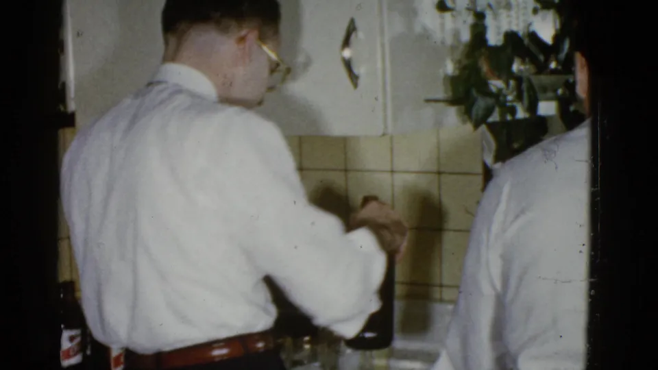 A 1951 Boston home-movie frame shows the man facing the kitchen counter as he handles a drink, with another adult partly cropped at right.