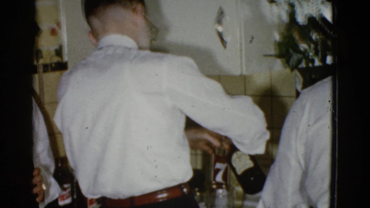 Two men in white shirts stand close together in a kitchen while one pours from a dark bottle.