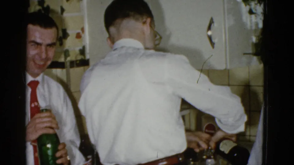 A young man in a red tie looks toward the camera with a green bottle as drinks are prepared in a 1951 Boston kitchen setting.