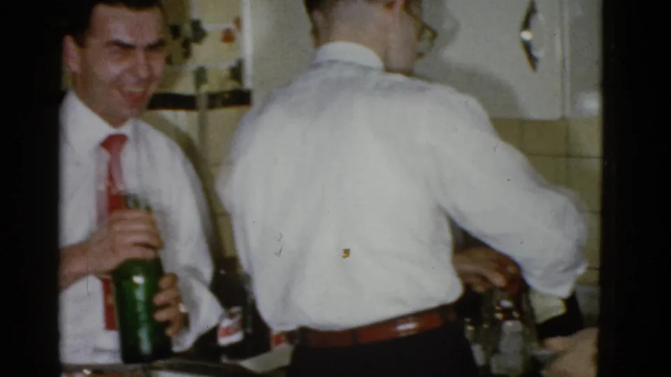 In a 1951 Boston private kitchen, one man works at the drinks while another smiles with a bottle in hand.
