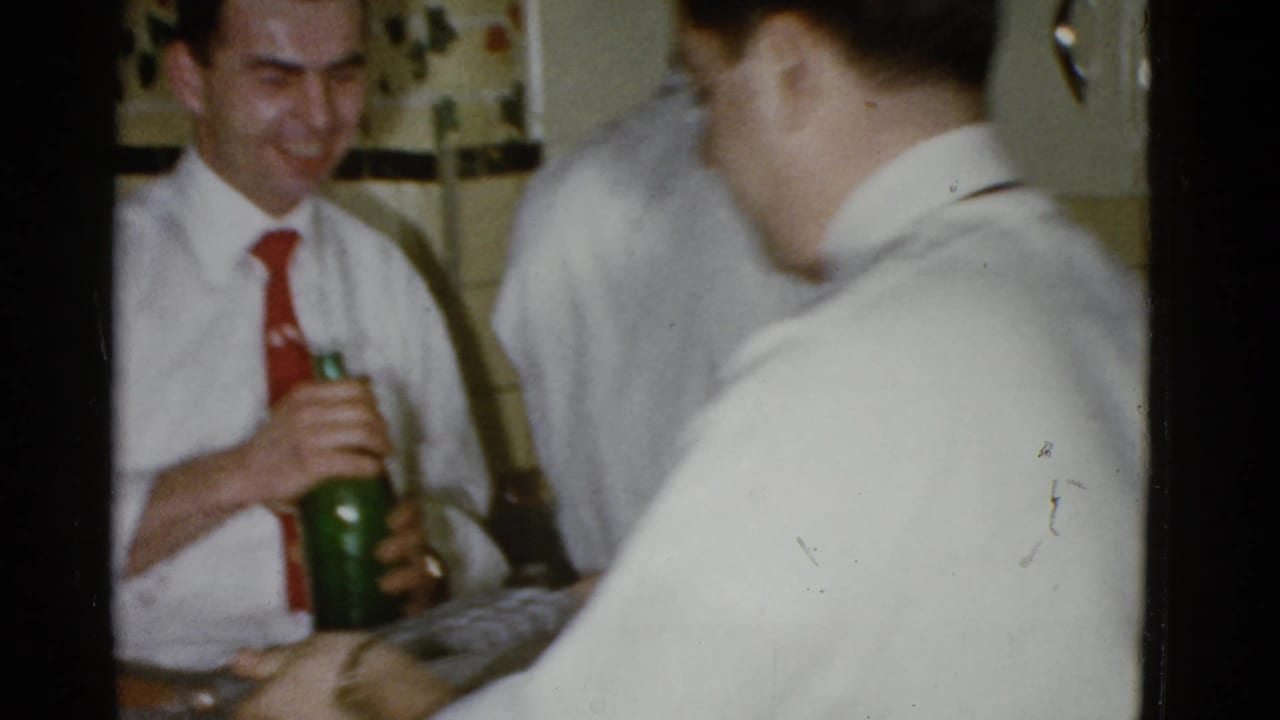 Man in a white shirt and red tie holding a green bottle in a kitchen while others stand nearby.