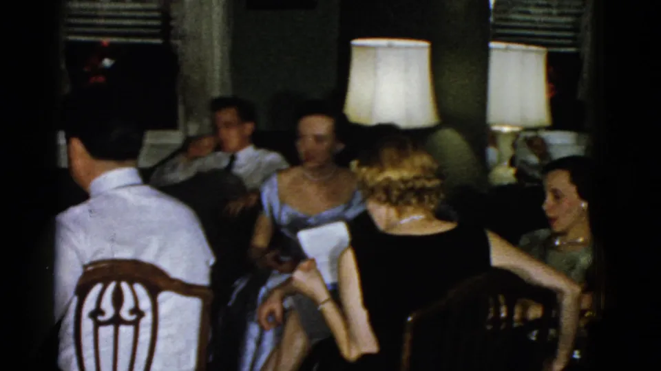 1951 Boston home-movie frame of adults in formal attire seated in a dim living room during a private social gathering.