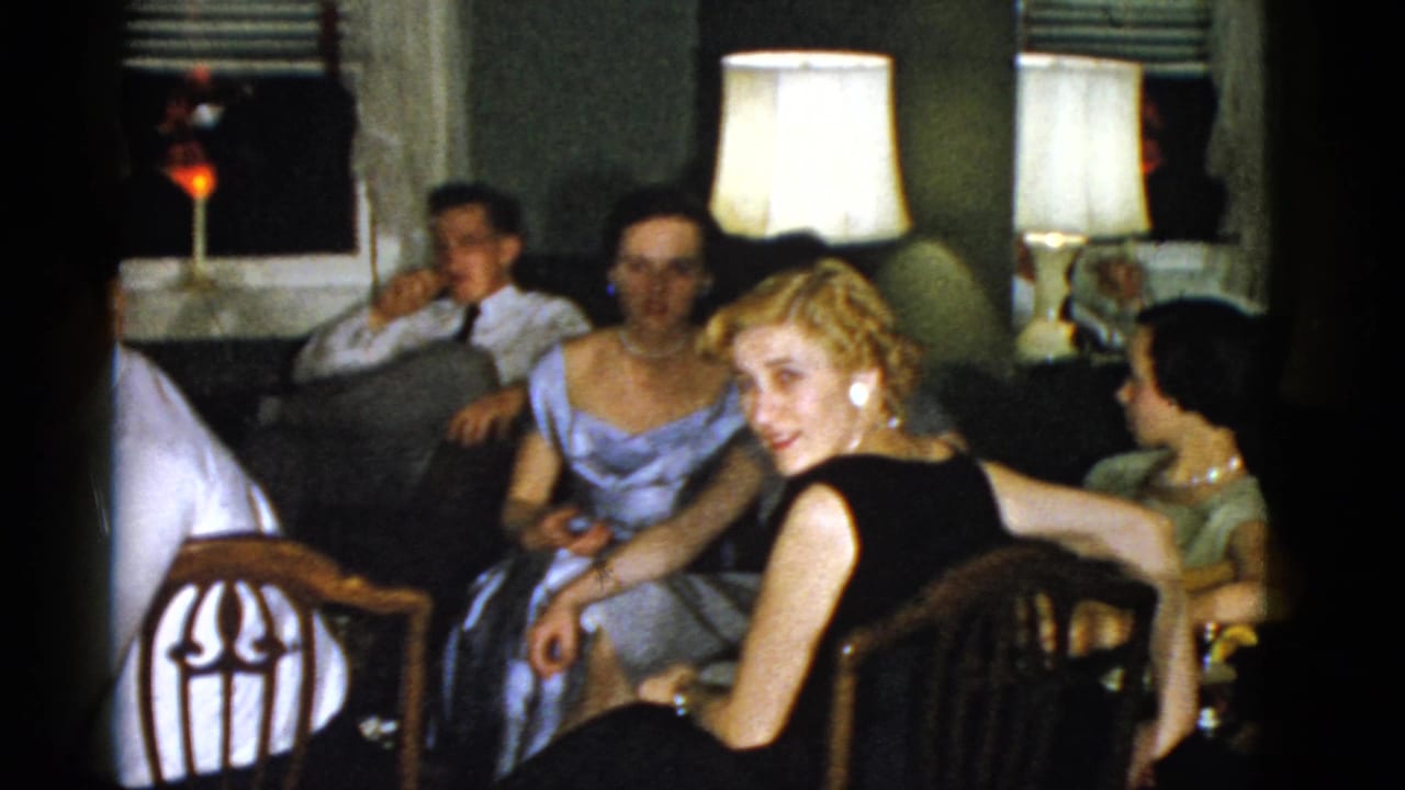 Adults in formal attire seated in a dim living room with table lamps and chairs.