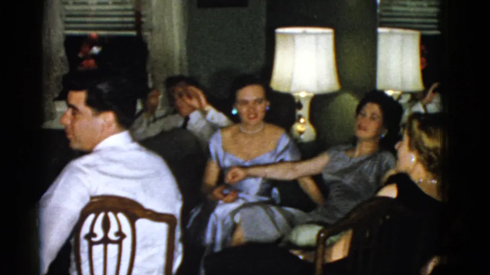 Boston 1951 frame with a man in a white shirt in the foreground and adults seated in formal attire around a dim living room.