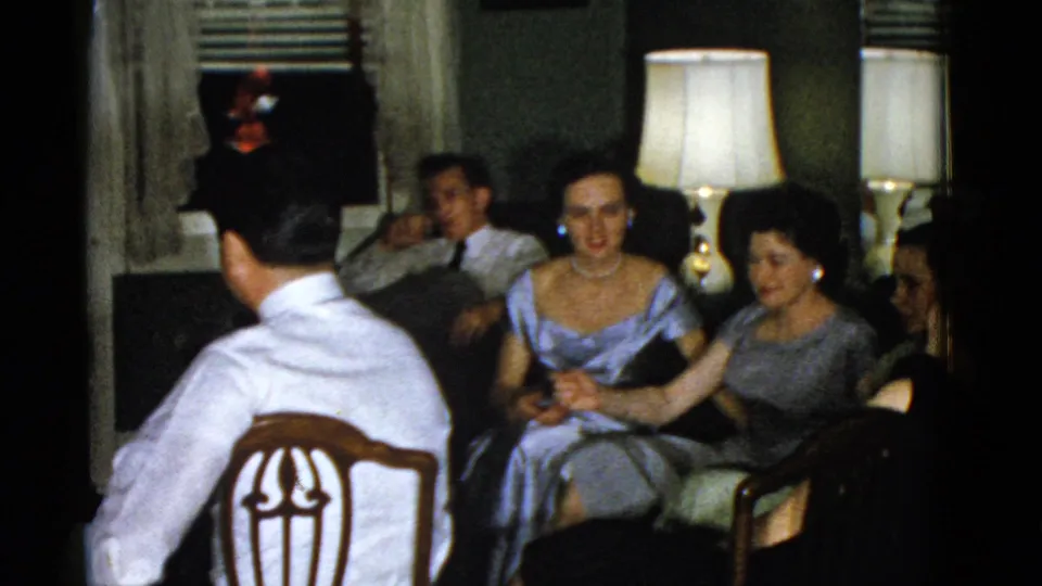 Final Boston 1951 home-movie frame showing adults seated near table lamps in a private living room, with one man viewed from behind.