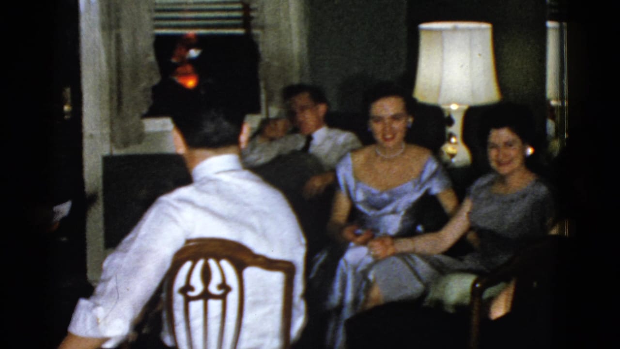 Adults in formal clothing seated in a dim living room while a man faces away in a wooden chair.