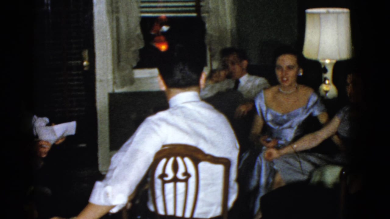 Seated adults in a dark living room, including a woman in a blue dress near a lit table lamp.