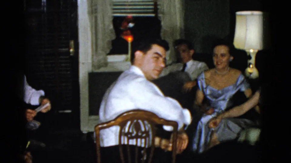 1951 Boston home-movie frame showing a man turning toward the camera while adults sit behind him in a dim domestic interior.