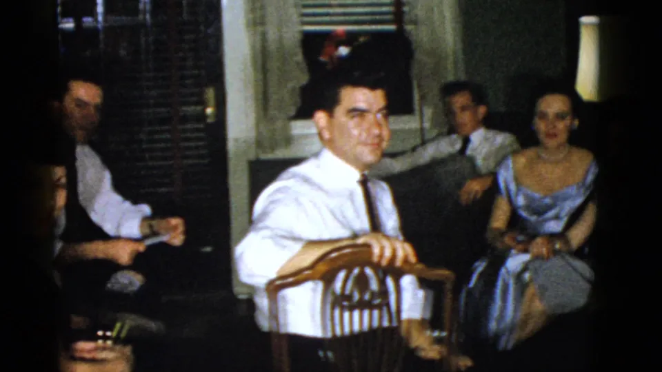 1951 Boston family gathering frame with a foreground man in a white shirt facing camera, seated adults, a blue satin dress, and warm lamp light.