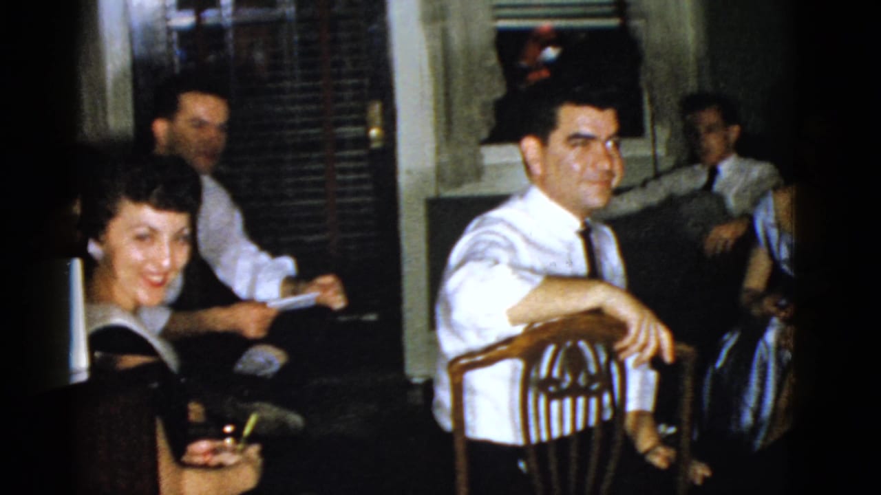 Adults seated in a dim home interior, with a woman smiling and a man leaning on a wooden chair.