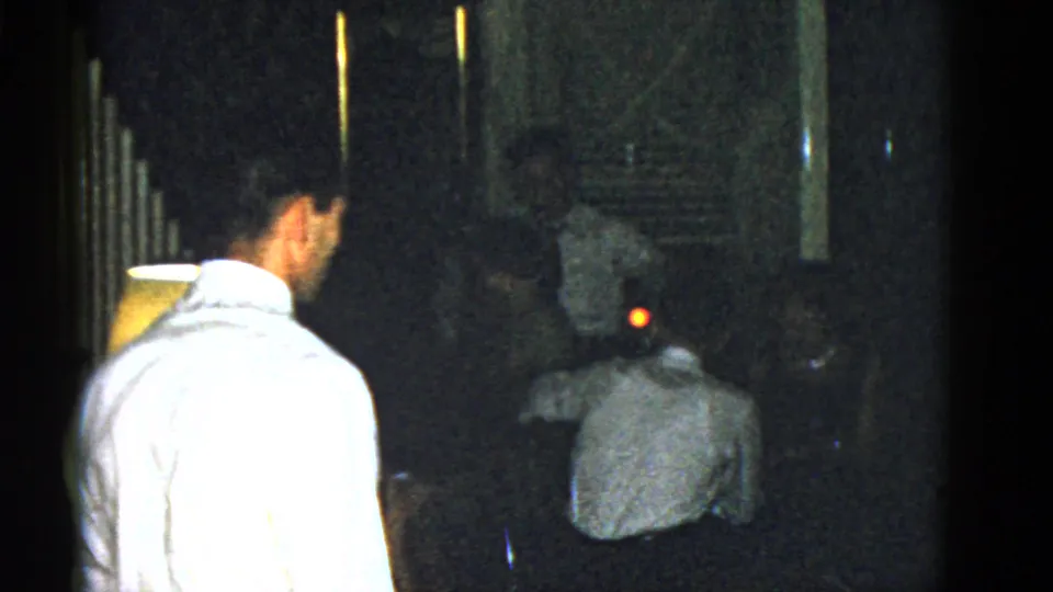 Dark Boston 1951 8mm frame with a man in a light shirt, shadowed figures, reflective glass, and a small red light.