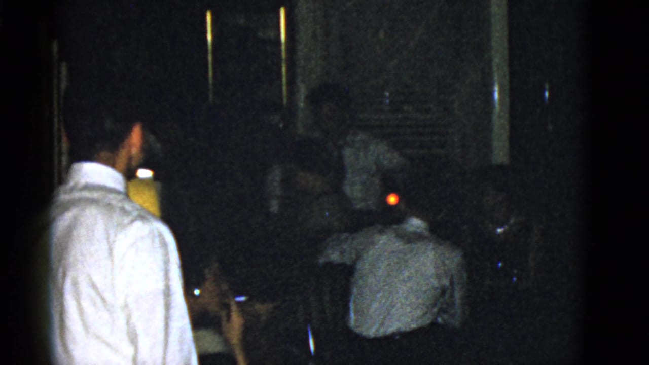 Dim room with a standing man in a white shirt near seated adults.