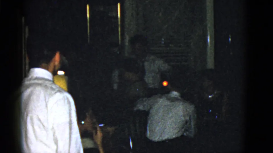 1951 Boston home-movie frame showing a man in a white dress shirt standing near seated adults in a dark private interior.