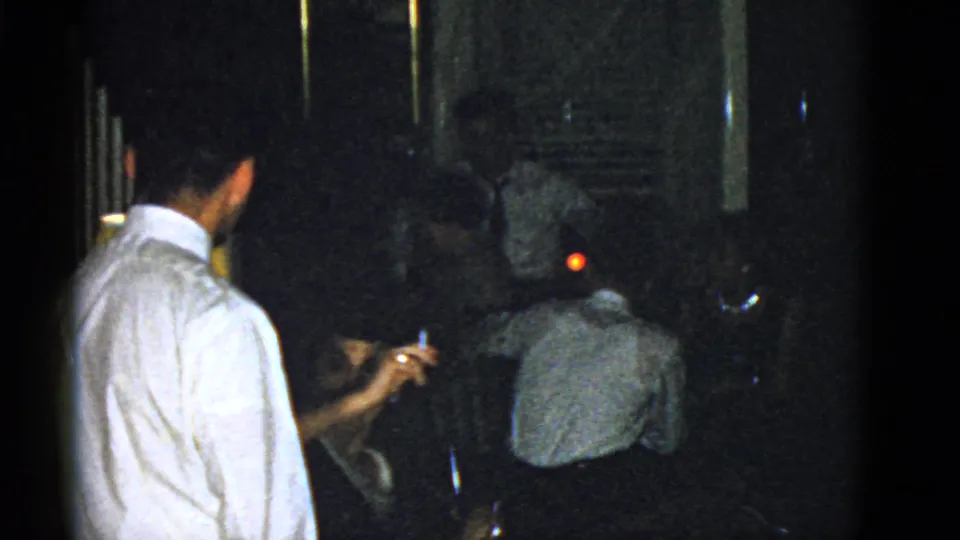 A 1951 Boston indoor home-movie frame with a standing man and seated adults conversing near a small red glow in the background.