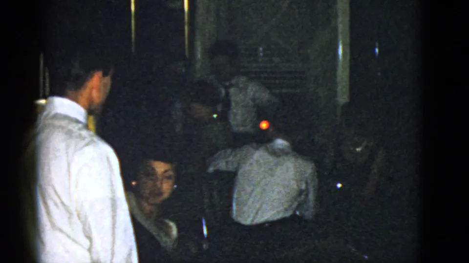 1951 Boston home-movie still with a seated woman partly visible and a man in a white shirt standing in a dim interior.